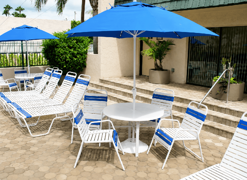 Commercial pool deck featuring white and royal blue vinyl strap dining chairs, round fiberglass tables, and royal blue Frankford market umbrellas creating a shaded outdoor dining area.